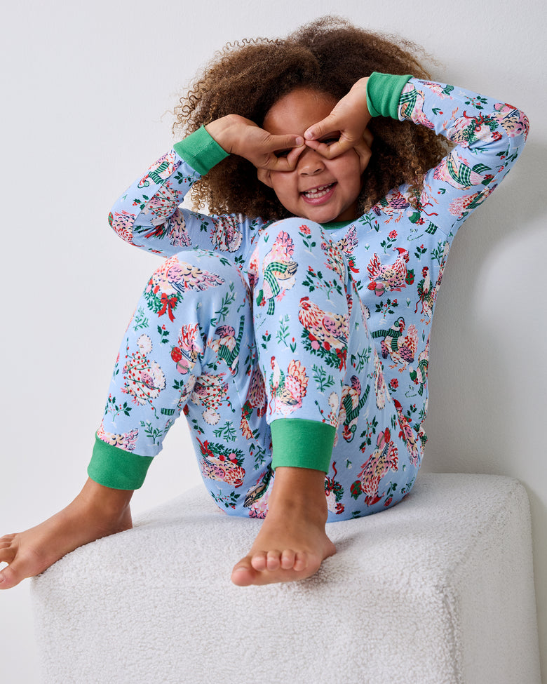 Child wearing a pale blue Holiday Hens pajama set sits on a white ottoman with both hands over their eyes.