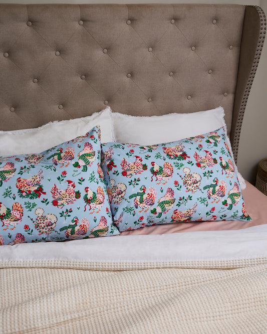 Two pale blue Holiday Hens pillowcases with illustrated holiday hens on a bed.