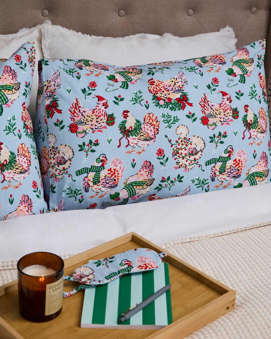 Two pale blue Holiday Hens pillowcases displayed on a bed beside a tray with a candle, notebook, and pen.