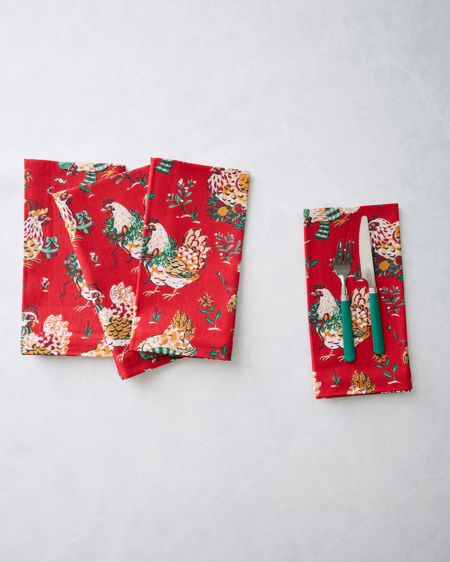 Red holiday hen-print dinner napkins arranged beside a napkin holding a fork and knife.
