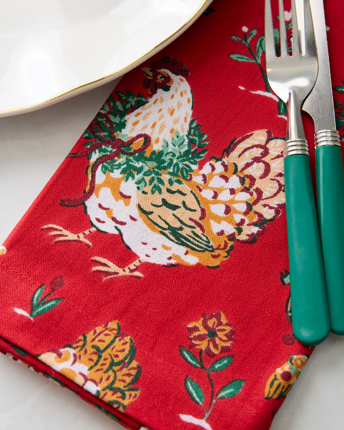 Close-up of a red holiday hen-print napkin beside a white plate and green-handled fork and knife.