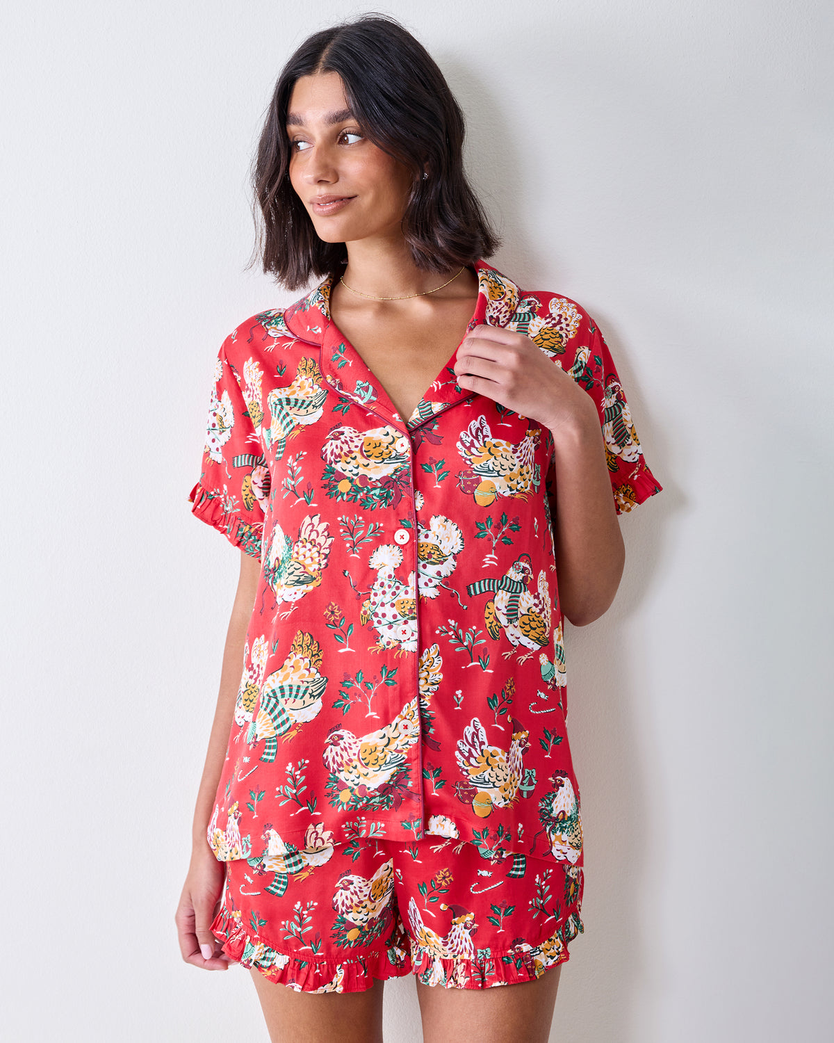 Front three-quarter view of a model wearing the red Holiday Hens short pajama set with ruffled trim and chicken print.