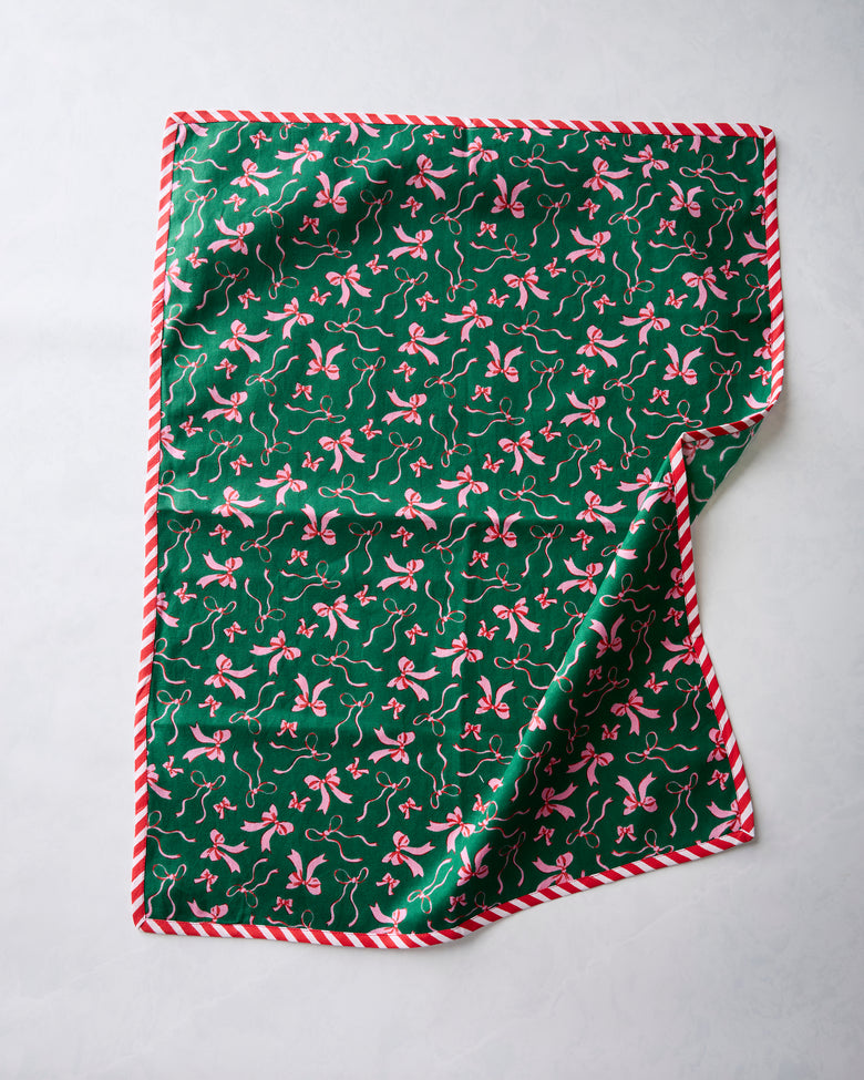 Green tea towel with a repeating pink bow print and red-and-white striped edging, shown with one corner folded back.