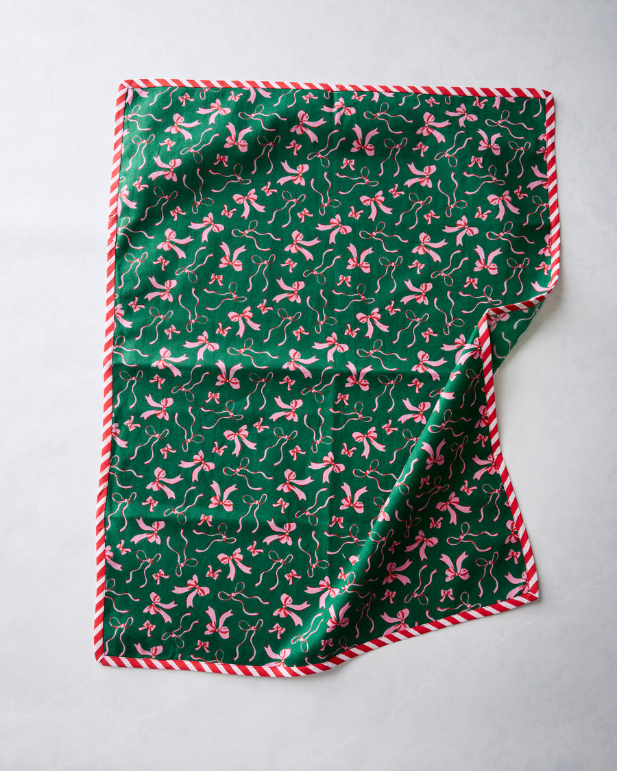 Green tea towel with a repeating pink bow print and red-and-white striped edging, shown with one corner folded back.