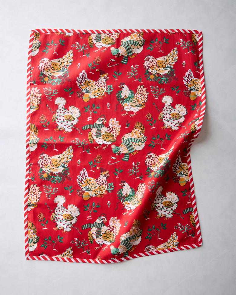 Red tea towel with a repeating illustrated holiday hen print and red-and-white striped edging, shown with one corner folded.