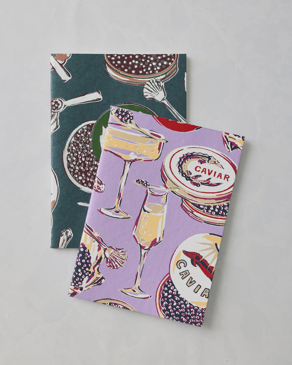 Two Champagne and Caviar notebook covers, lavender and dark teal, overlapping to show their champagne and caviar illustrations; “CAVIAR” is visible on the lavender cover.