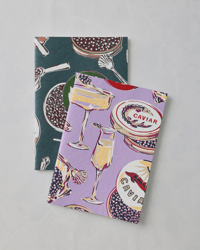 Two Champagne and Caviar notebook covers, lavender and dark teal, overlapping to show their champagne and caviar illustrations; “CAVIAR” is visible on the lavender cover.