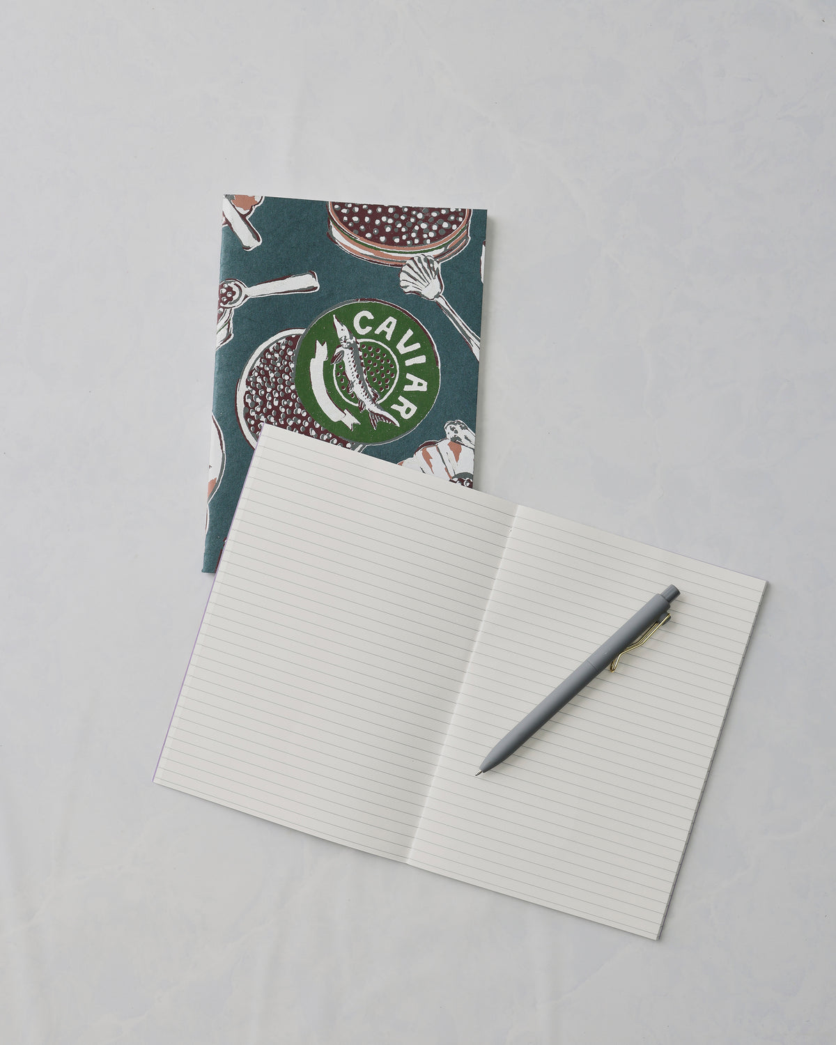 Champagne and caviar notebook set with a dark teal patterned notebook behind an open lined notebook and gray pen; the cover shows the word “CAVIAR.”
