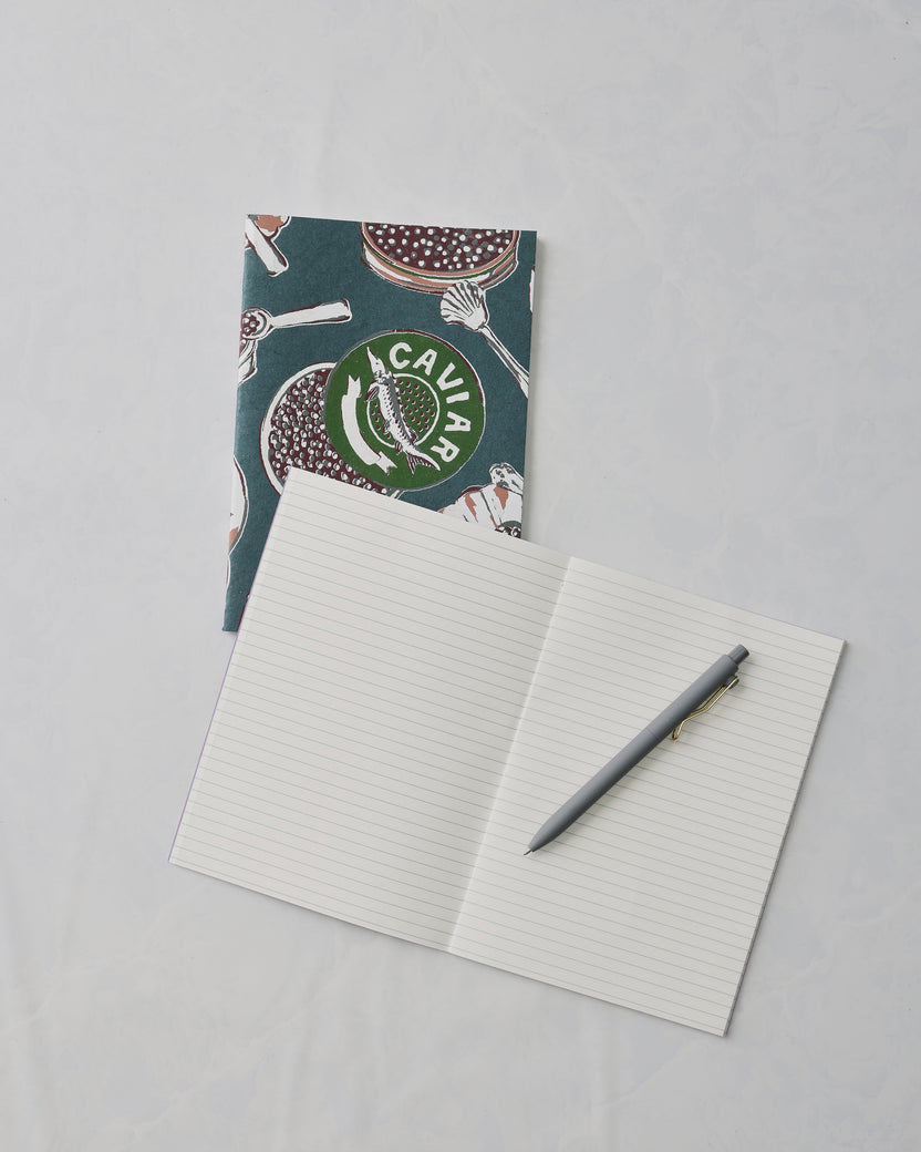 Champagne and caviar notebook set with a dark teal patterned notebook behind an open lined notebook and gray pen; the cover shows the word “CAVIAR.”