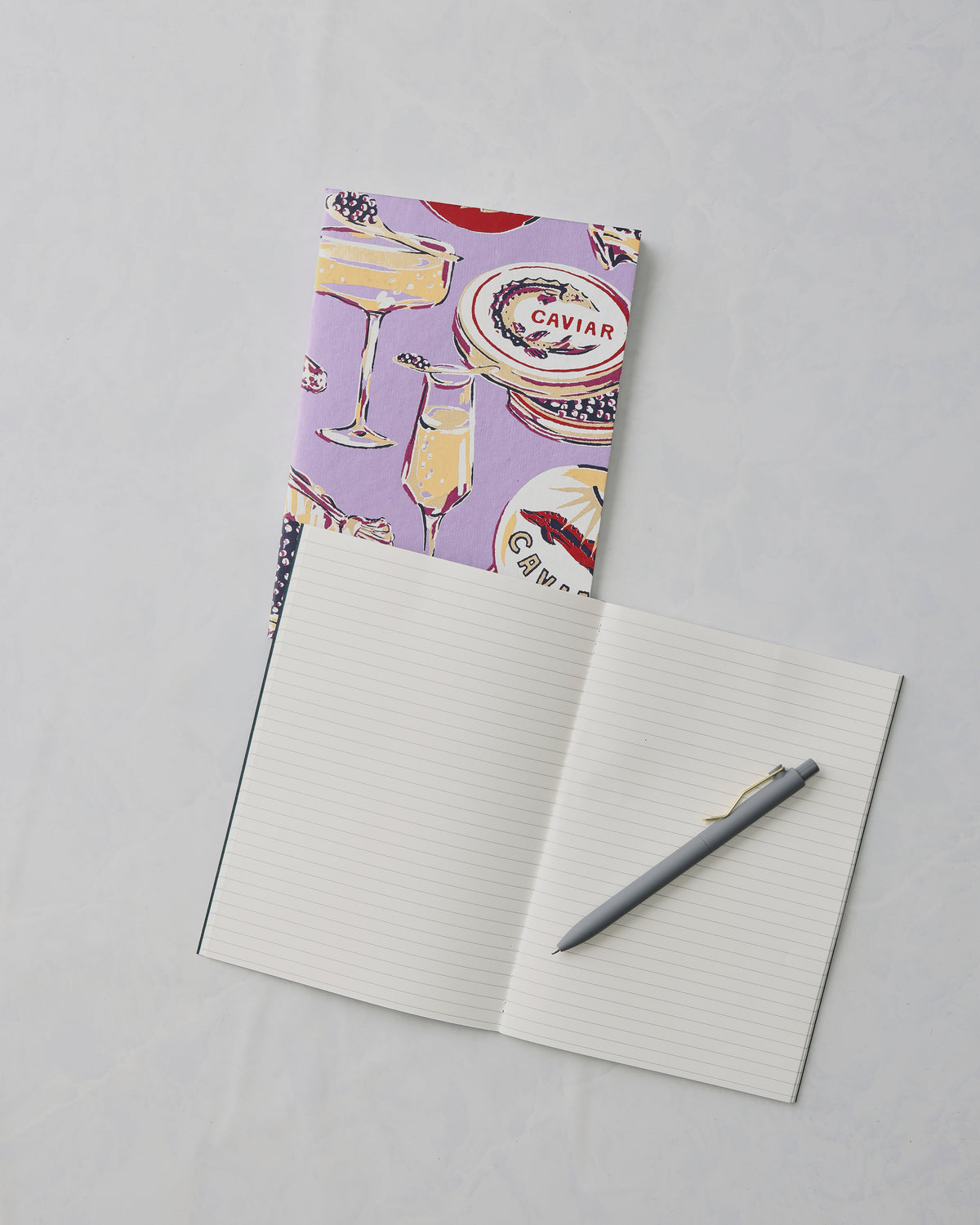 Lavender Champagne and Caviar notebook behind an open lined notebook with a gray pen; the cover shows champagne and caviar illustrations and the word “CAVIAR.”