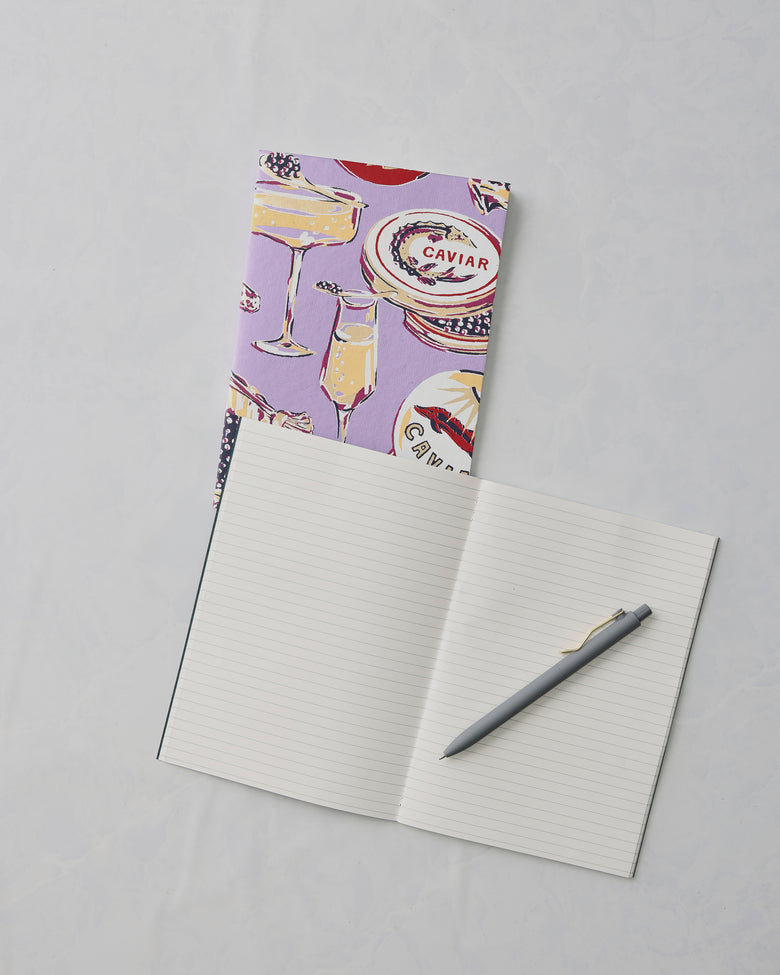 Lavender Champagne and Caviar notebook behind an open lined notebook with a gray pen; the cover shows champagne and caviar illustrations and the word “CAVIAR.”