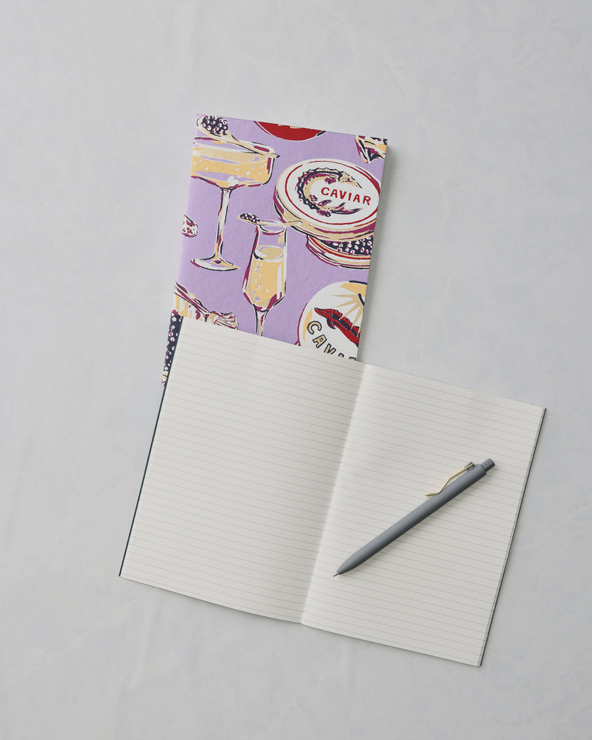Lavender Champagne and Caviar notebook behind an open lined notebook with a gray pen; the cover shows champagne and caviar illustrations and the word “CAVIAR.”