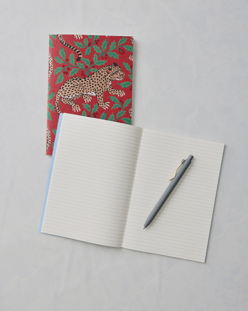 Open lined notebook with a gray pen, beside a closed red notebook with leopard and leafy print.