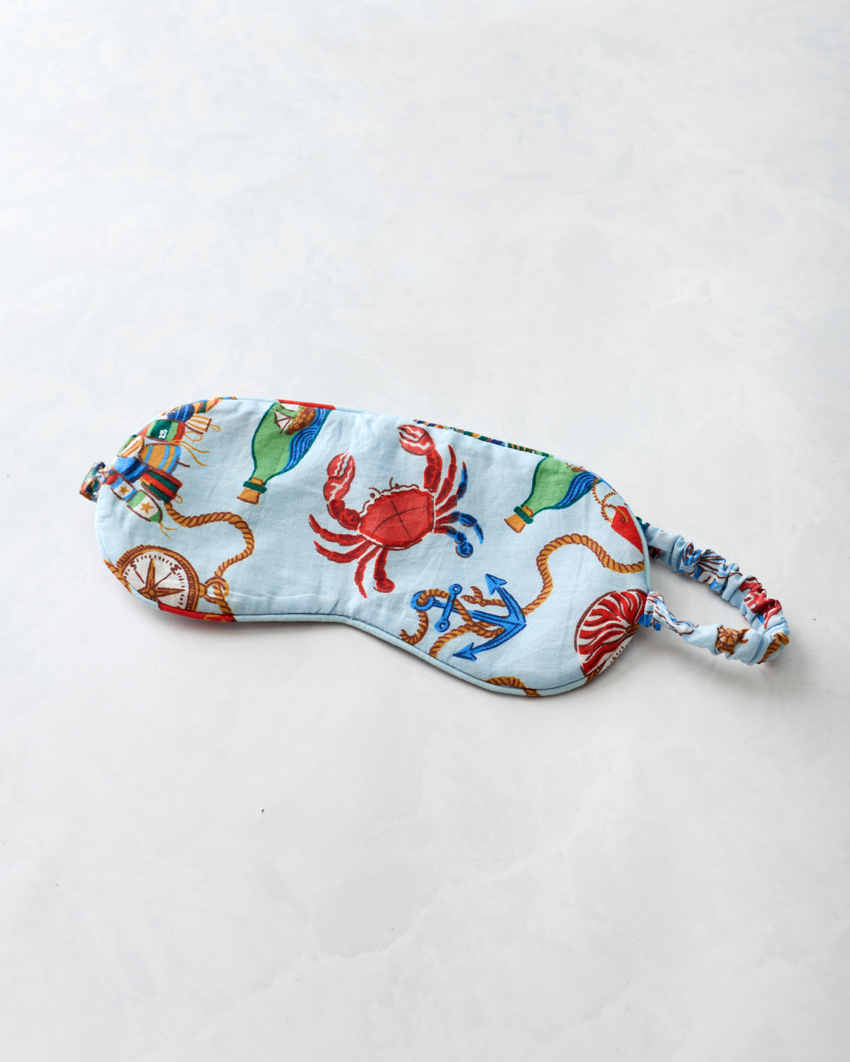 Blue sleep eye mask with a nautical print featuring a red crab, anchor, seashell, compass, and rope motifs.