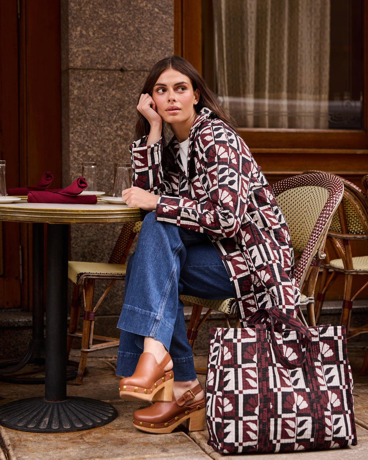 Person seated at a café wearing a burgundy, cream, and black geometric-patterned jacket beside a matching large tote bag.