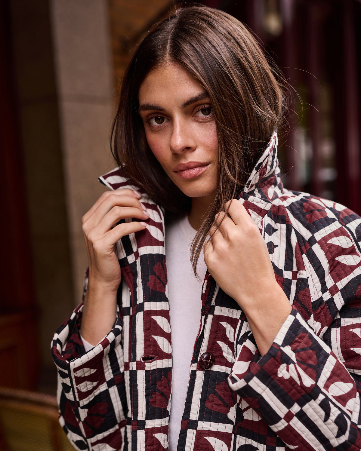 Close portrait view of a person holding the collar of a burgundy, cream, and black geometric-patterned Spencer jacket.