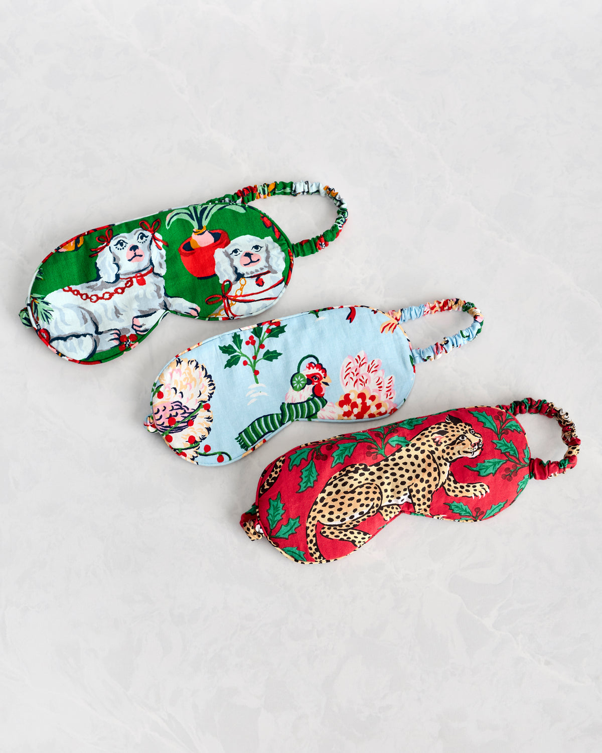 Three printed sleep masks arranged diagonally: green with dogs, light blue with holiday foliage and birds, and red with a leopard.