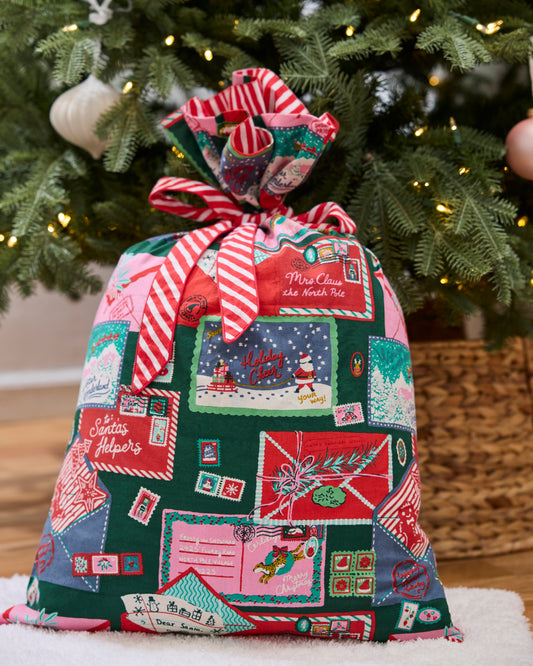 Christmas gift sack tied with a red-and-white striped fabric bow, decorated with illustrated holiday envelopes and postage stamps in front of a lit tree.