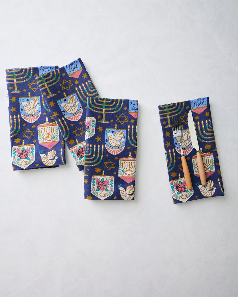 Four folded Hanukkah blue dinner napkins with menorahs, dreidels, stars, and dove motifs; one napkin holds a fork and knife.