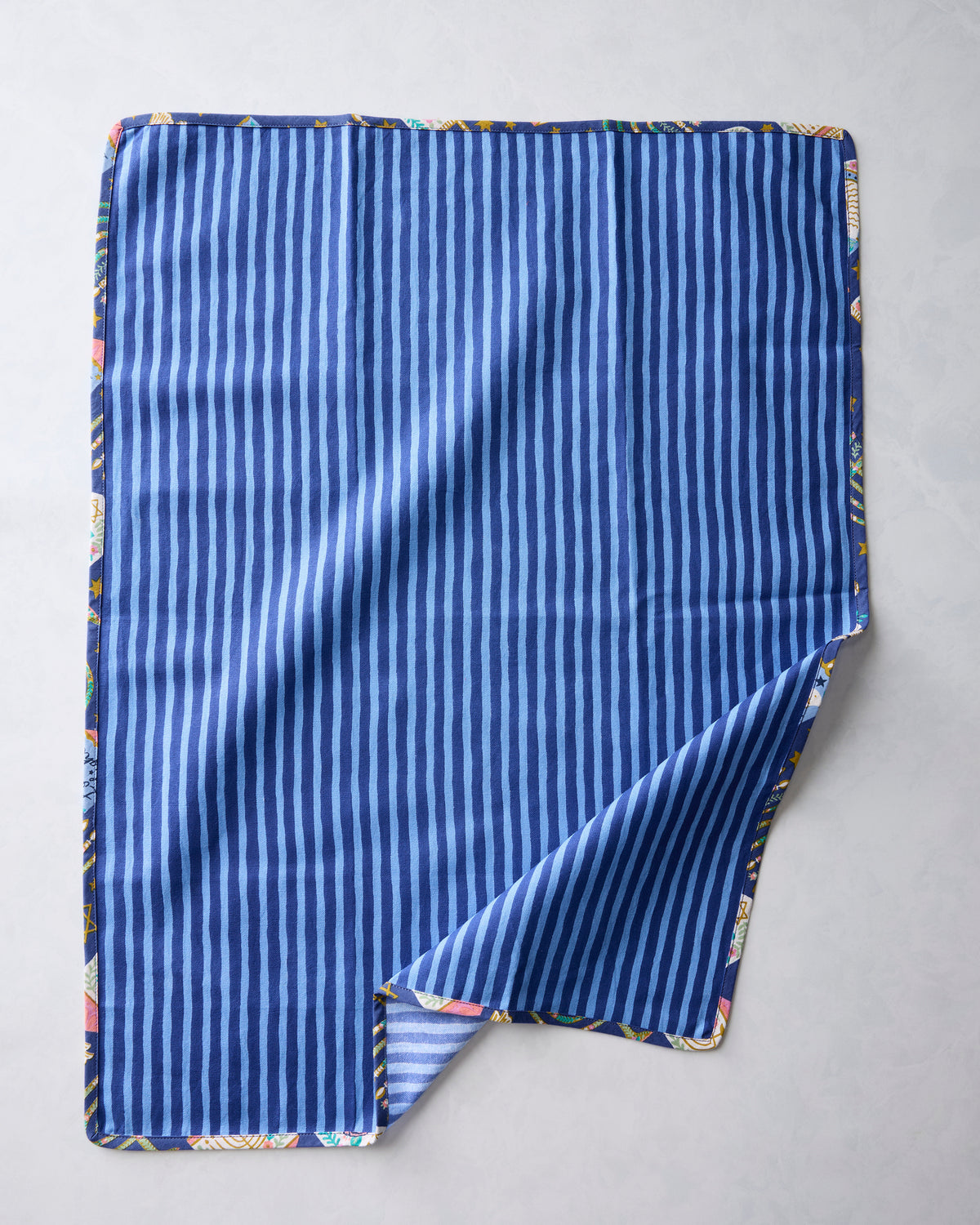 Blue tea towel showing a vertical blue stripe pattern, with a folded corner revealing the printed Hanukkah design along the edge.