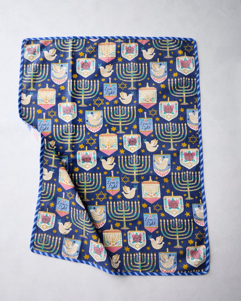 Blue tea towel with repeating Hanukkah-themed symbols, including menorahs, stars, dreidels, and candles, with a striped border.