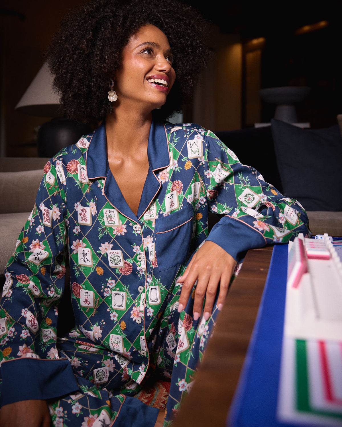 Person seated beside a mahjong table wearing the Mahj Garden Soho set with a patterned button-front top and matching pants.