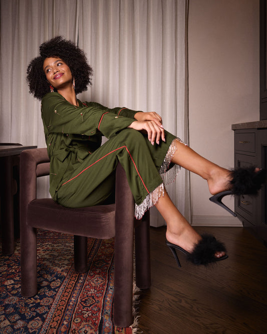 Seated view of the olive Matchmaker pajama set showing the belted top, piped pants, beaded tassel hems, and black feather-trimmed heeled shoes.