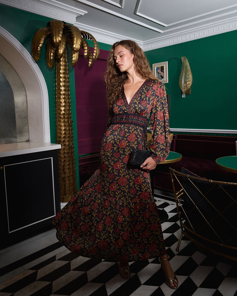 Person wearing a dark floral long-sleeve dress and holding a black clutch in an interior with green walls.
