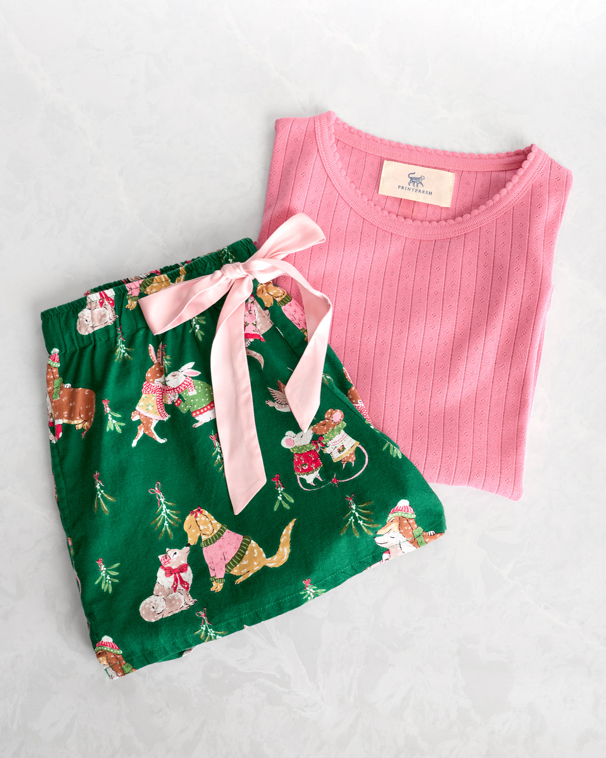 Pink long-sleeve top and green pajama shorts with holiday animal print, arranged with a pale pink ribbon tied between them.