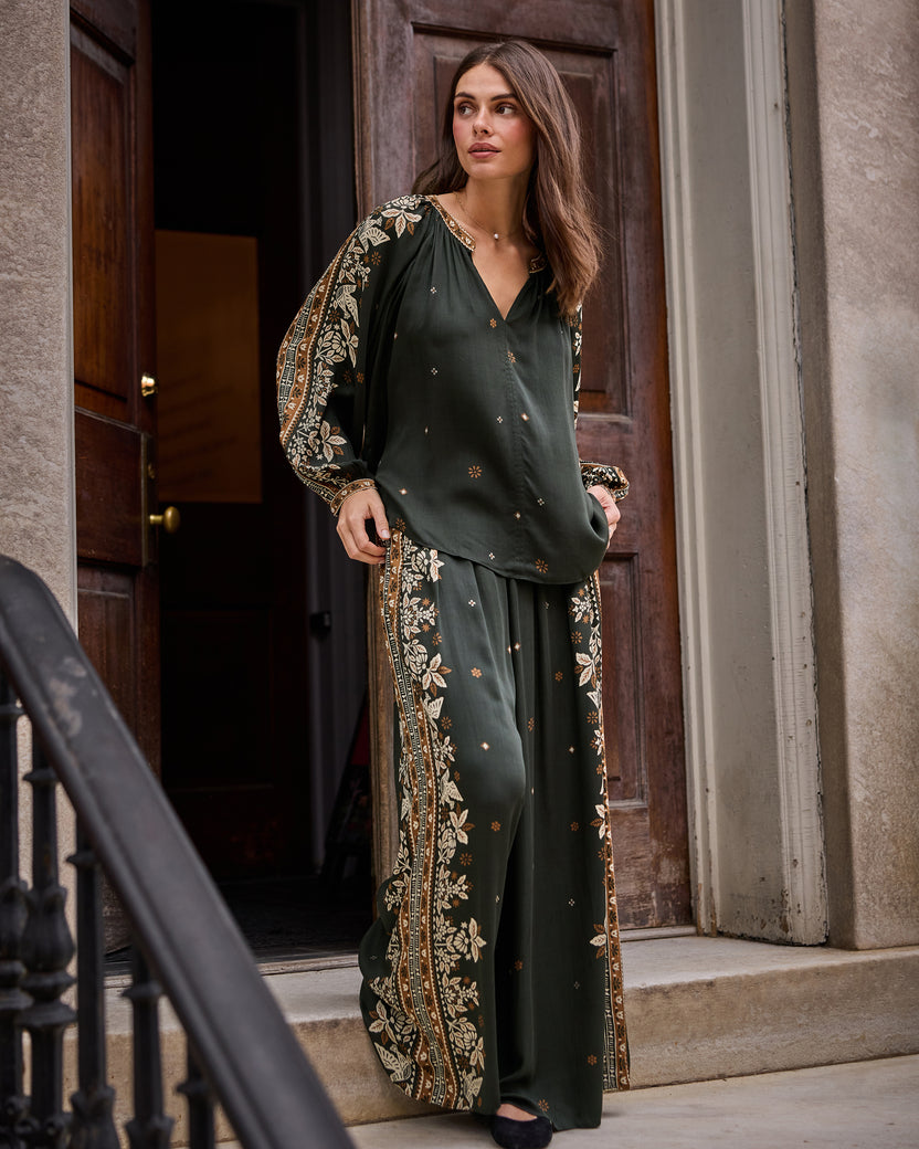Full-length front view of the Hallie Blouse and Sasha Pants in dark green with floral patterned side panels, standing in a doorway.