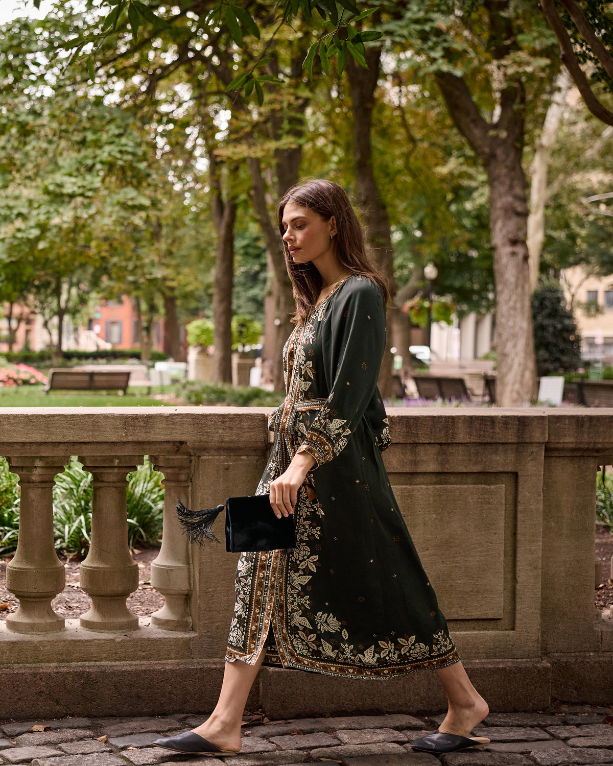 Side view of a person wearing the Hunter Dress in dark green with floral patterned borders, walking outdoors while carrying a black clutch.