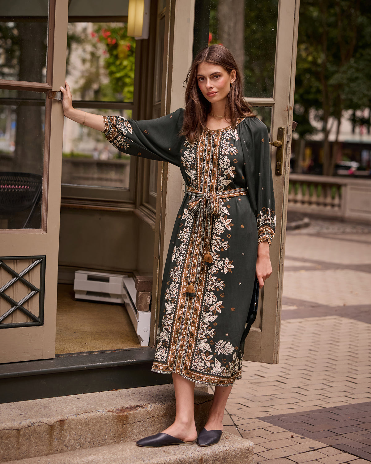 Person wearing the Solstice Tapestry Hunter dress in a doorway, showing its tie waist, long sleeves, side slit, and patterned front panel.