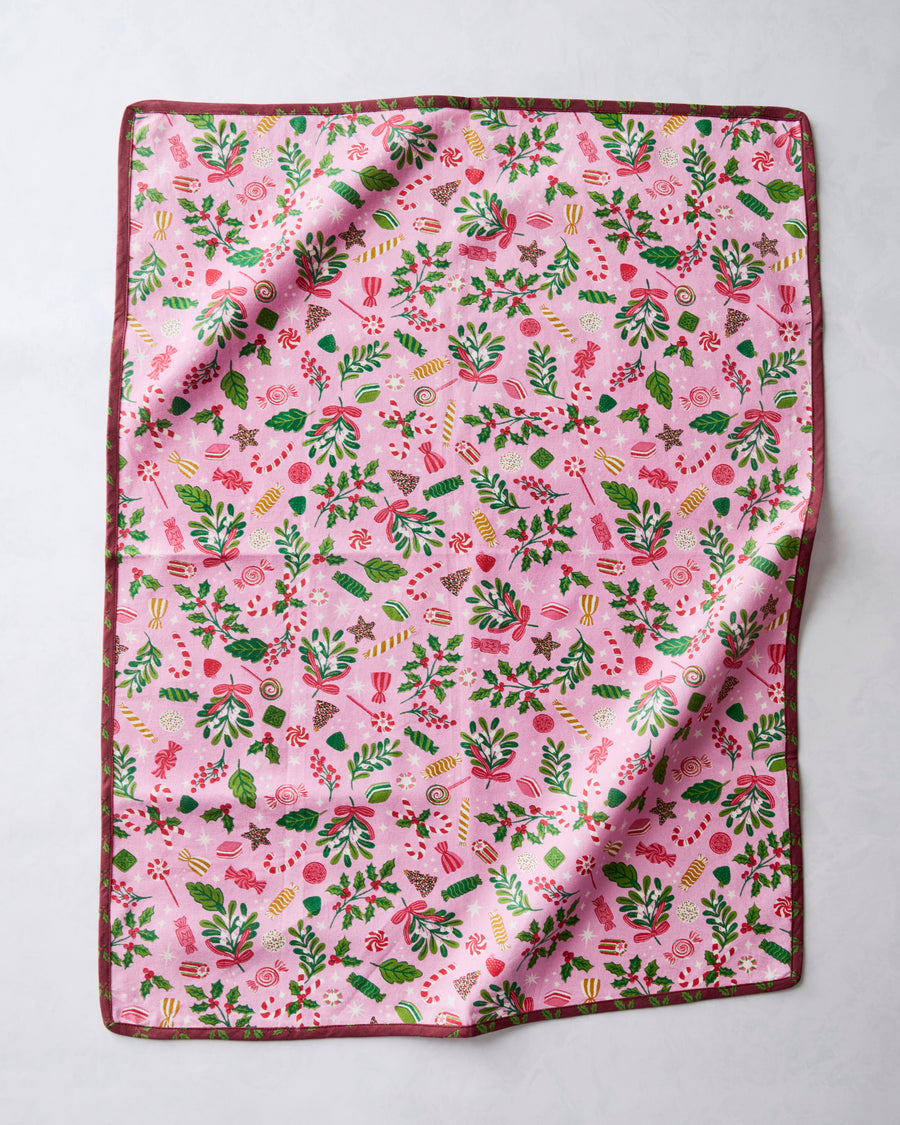 Tea towel with a pink holiday candy, holly, and foliage print, shown flat with a folded corner.