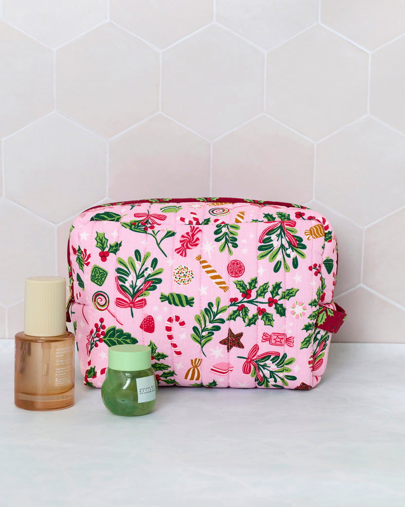 Candy pink quilted pouch with a colorful holiday print, shown upright beside two cosmetic containers for scale.