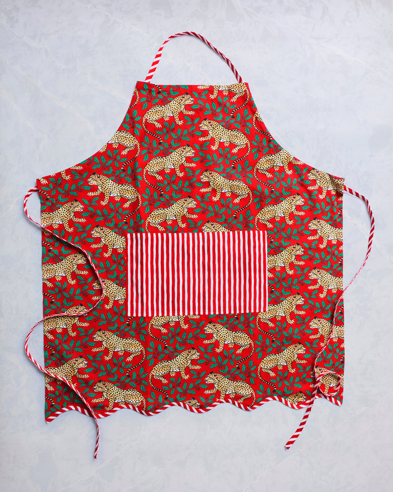 Red Winter Holly Bagheera apron laid flat, with spotted leopards and holly leaves, striped neck and waist ties, and a front pocket.