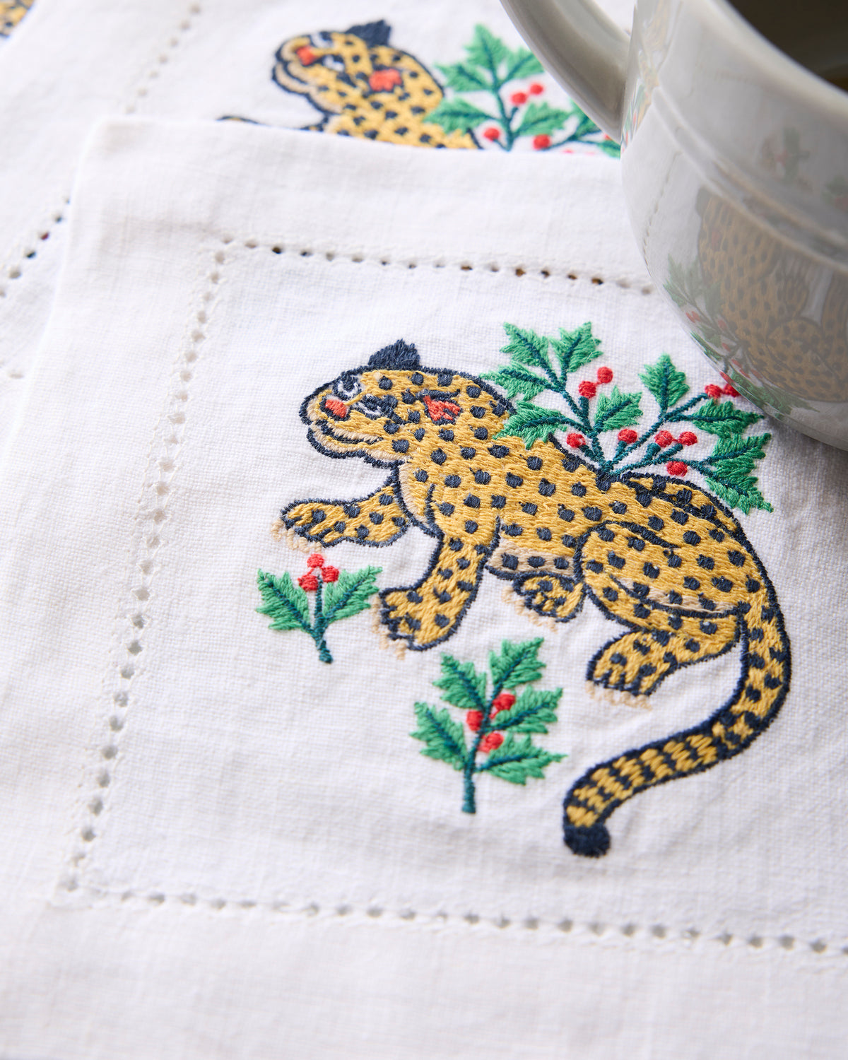 Close-up of an embroidered Winter Holly Bagheera cocktail napkin showing a spotted cat surrounded by holly branches and red berries.