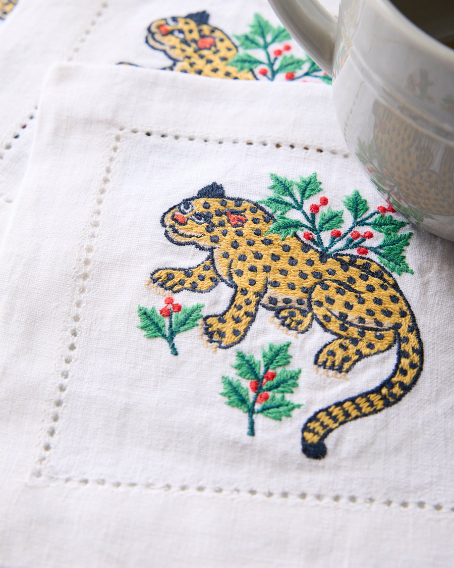 Close-up of an embroidered Winter Holly Bagheera cocktail napkin showing a spotted cat surrounded by holly branches and red berries.