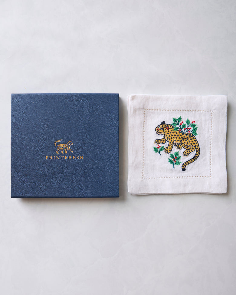 Winter Holly Bagheera cocktail napkin beside its blue Printfresh box, showing the embroidered spotted cat and holly design.