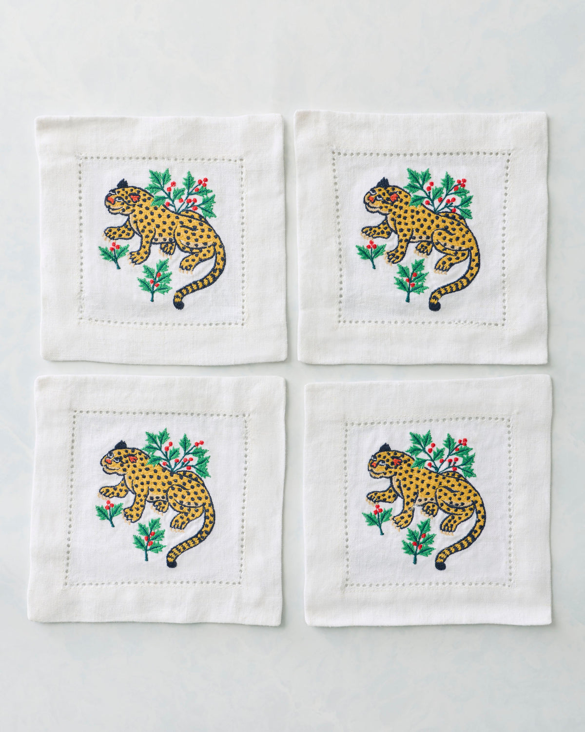 Four white cocktail napkins arranged in a square, each embroidered with a spotted yellow leopard and holly branches.