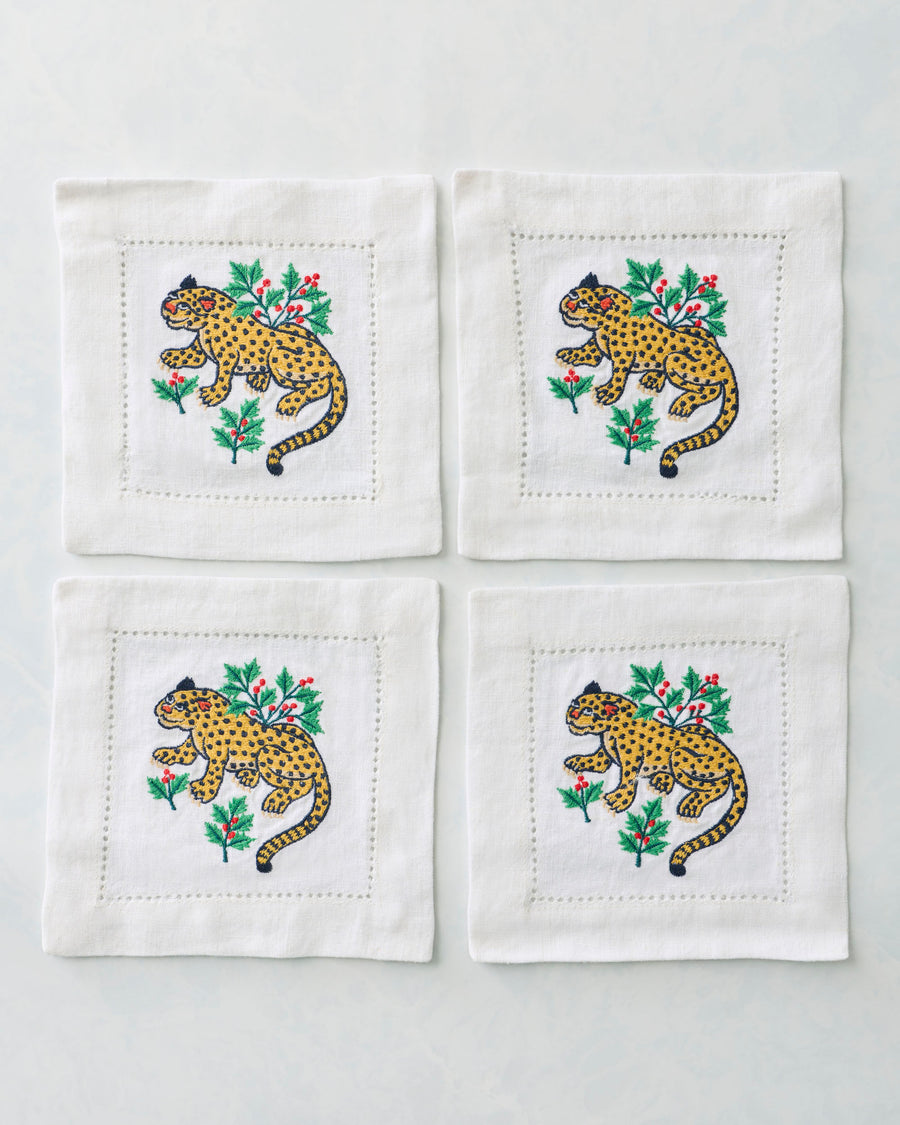 Four white cocktail napkins arranged in a square, each embroidered with a spotted yellow leopard and holly branches.