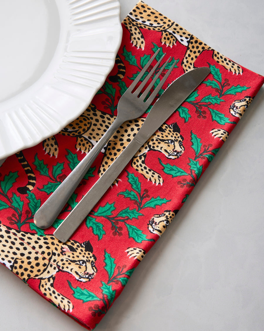 Ruby printed dinner napkin with leopard and holly motifs beneath a fork and knife beside a white plate.