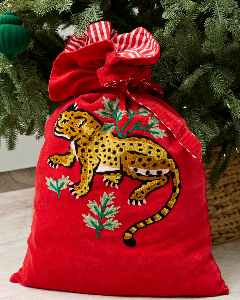 Red Winter Holly Bagheera gift sack with a striped drawstring tie and a leopard decoration surrounded by greenery, displayed beneath a Christmas tree.