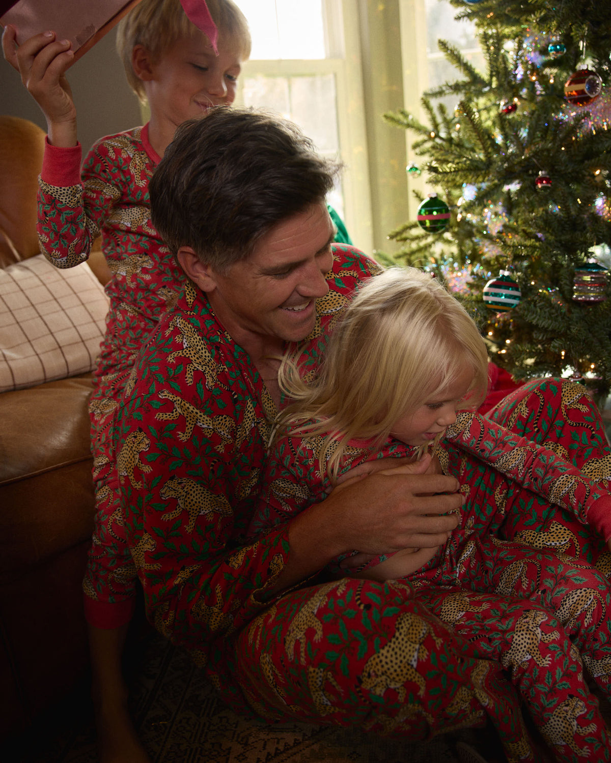 Three people wearing matching red leopard-and-holly pajamas beside a decorated Christmas tree.