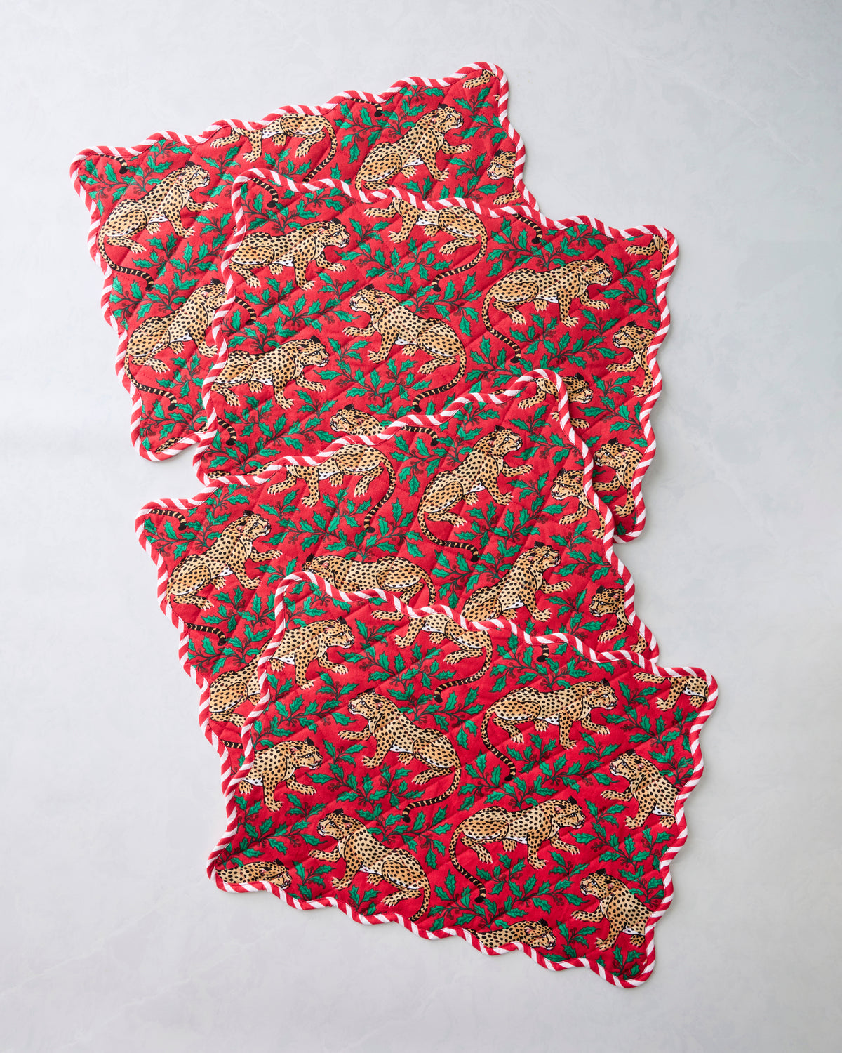Four Winter Holly Bagheera placemats overlapped in a diagonal arrangement, showing scalloped edges and a red leopard-and-holly print.