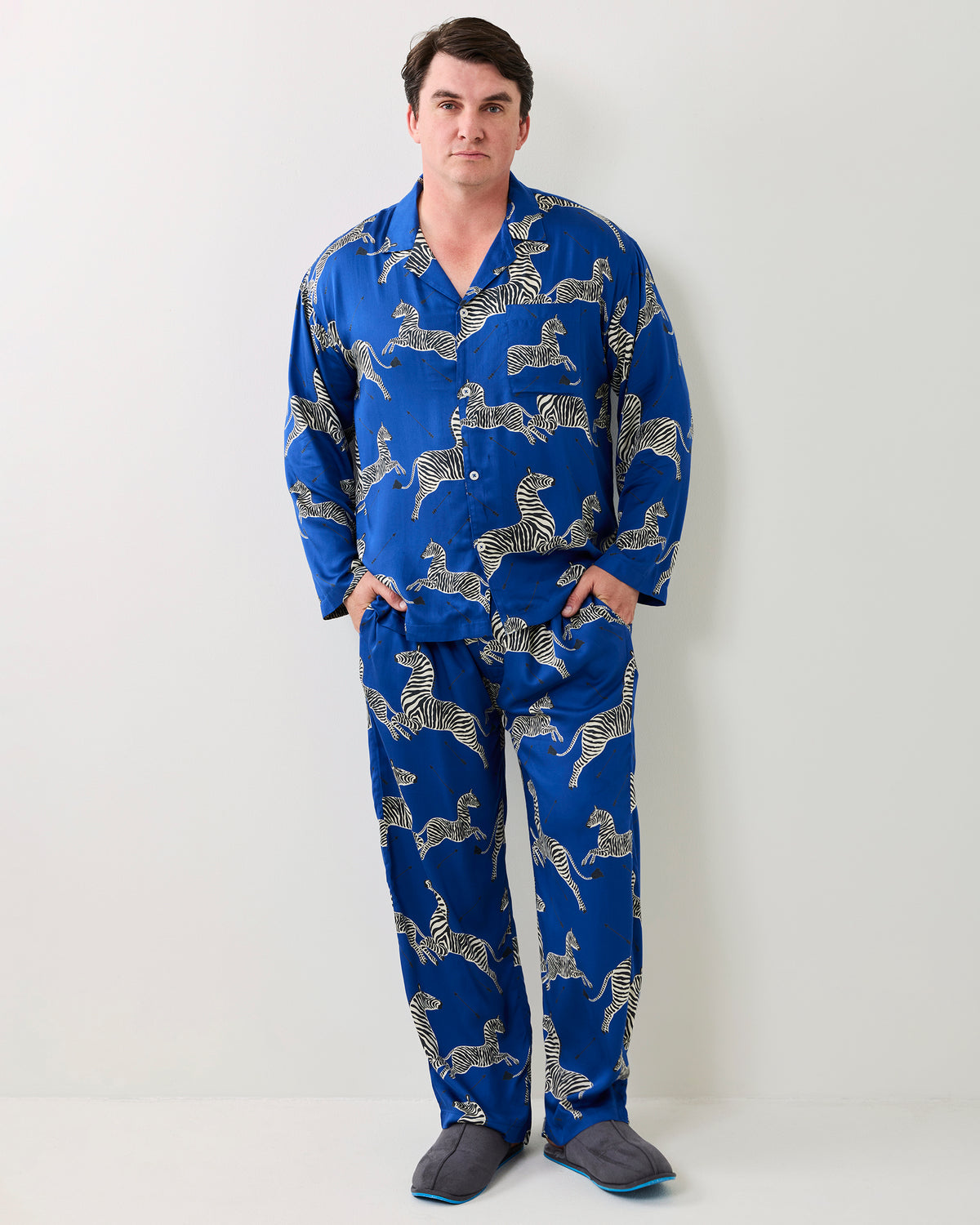 Adult model standing in a blue button-front pajama set with white zebra prints and slippers.