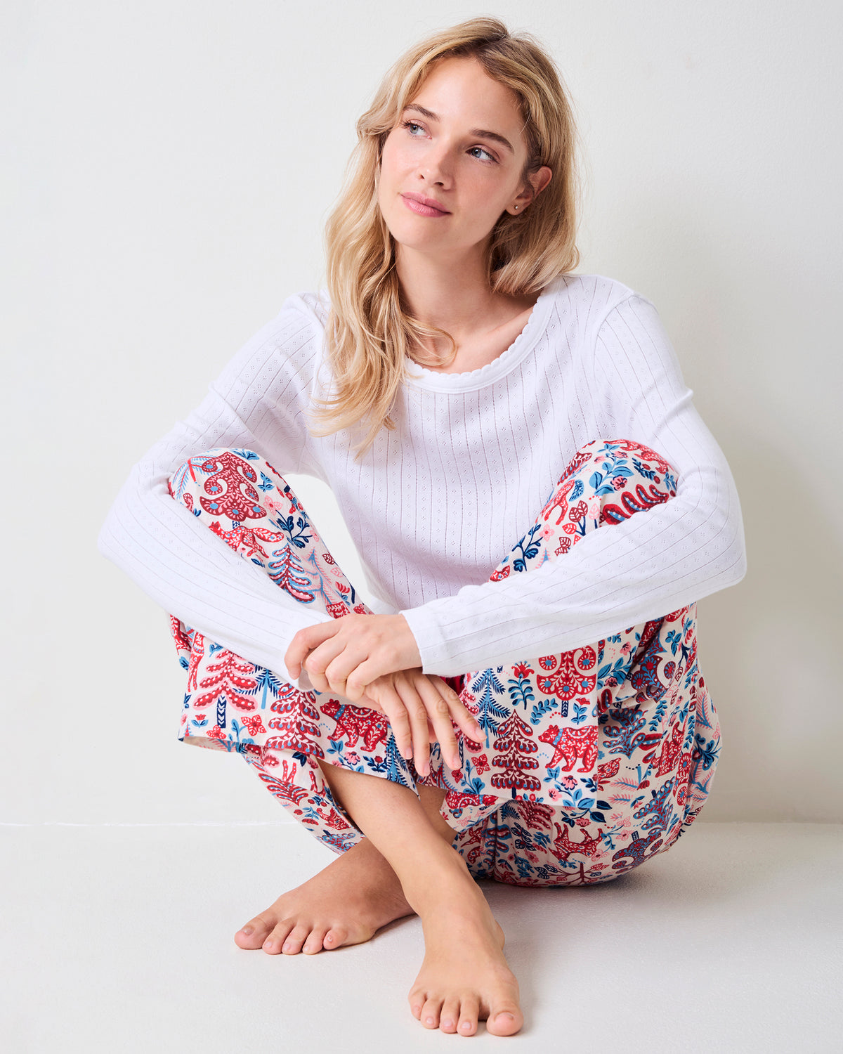 Adult model seated wearing cream Folklore Forest printed pajama pants with a white sweater.