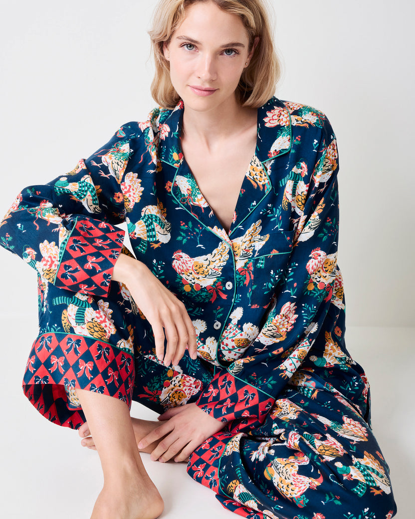 Seated model wearing a navy pajama set with festive hen print, white piping, and red patterned cuffs.