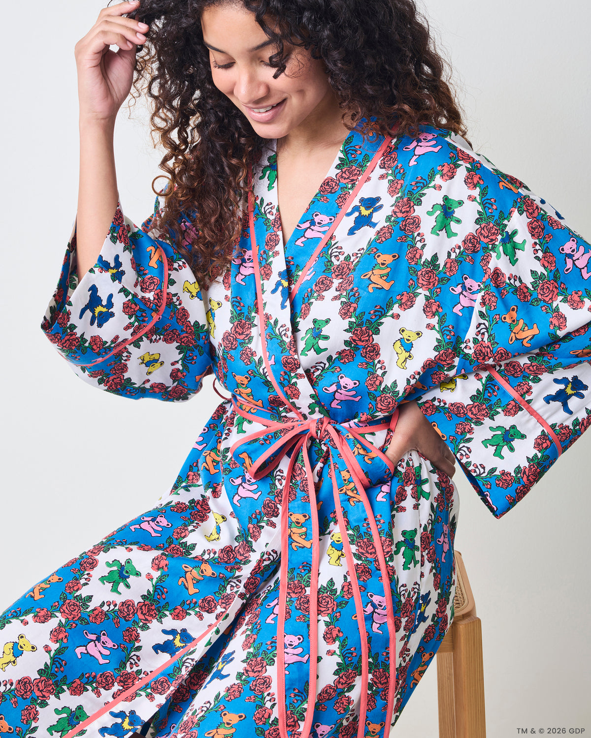 Angled seated view of a long robe with wide sleeves, pink trim, waist ties, and a blue-and-white bear-and-rose print.