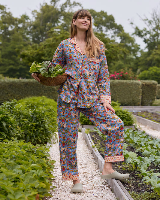 Full view of the Delphinium Blue Bee Tapestry tall long pajama set in a garden, with the wearer carrying a basket of leafy vegetables.