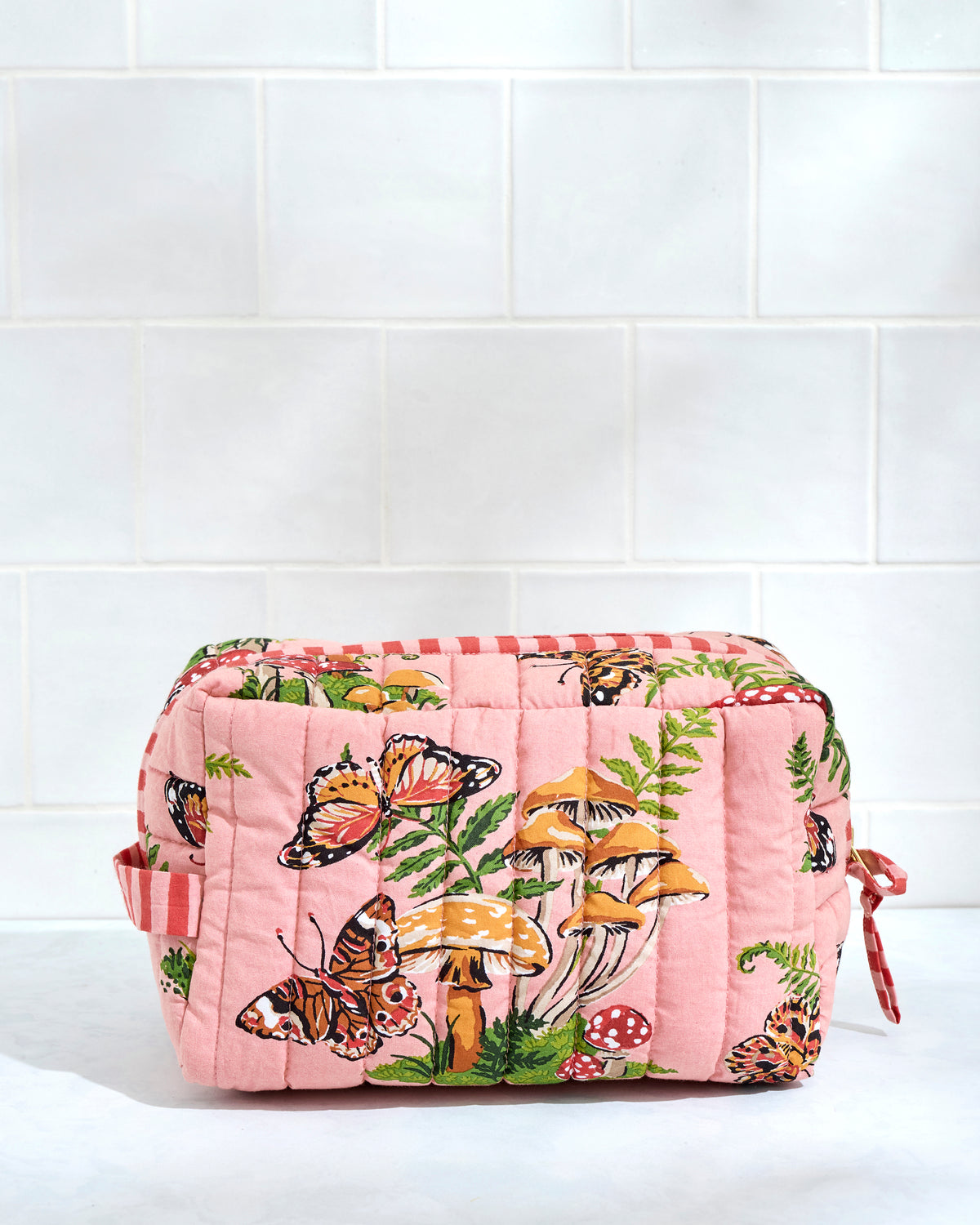 Front view of a quilted butterfly-print pouch with mushrooms and ferns, positioned against a white tile wall.